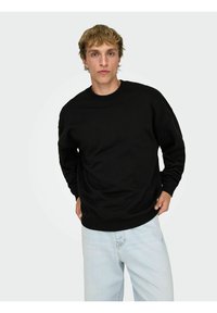 Felpa nera girocollo in materiale morbido con polsini e orlo a costine, abbinata a jeans azzurri. Il modello è in piedi davanti a uno sfondo semplice.