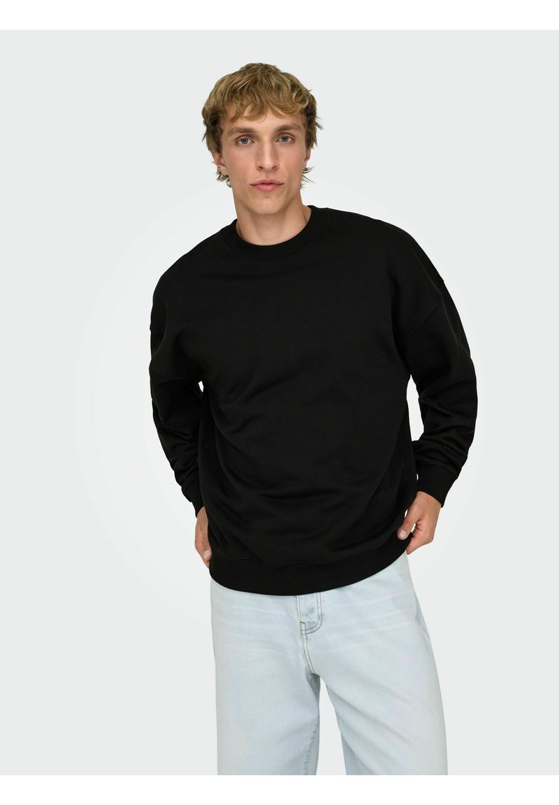 Felpa nera girocollo in materiale morbido con polsini e orlo a costine, abbinata a jeans azzurri. Il modello è in piedi davanti a uno sfondo semplice.