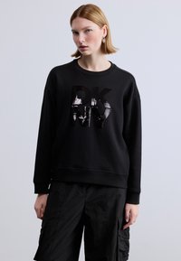 Μαύρο φούτερ με στρογγυλή λαιμόκοψη και υφή βρόχου, με γυαλιστερό μαύρο λογότυπο. Συνδυασμένο με φαρδιά μαύρη εργασιακή παντελόνα.