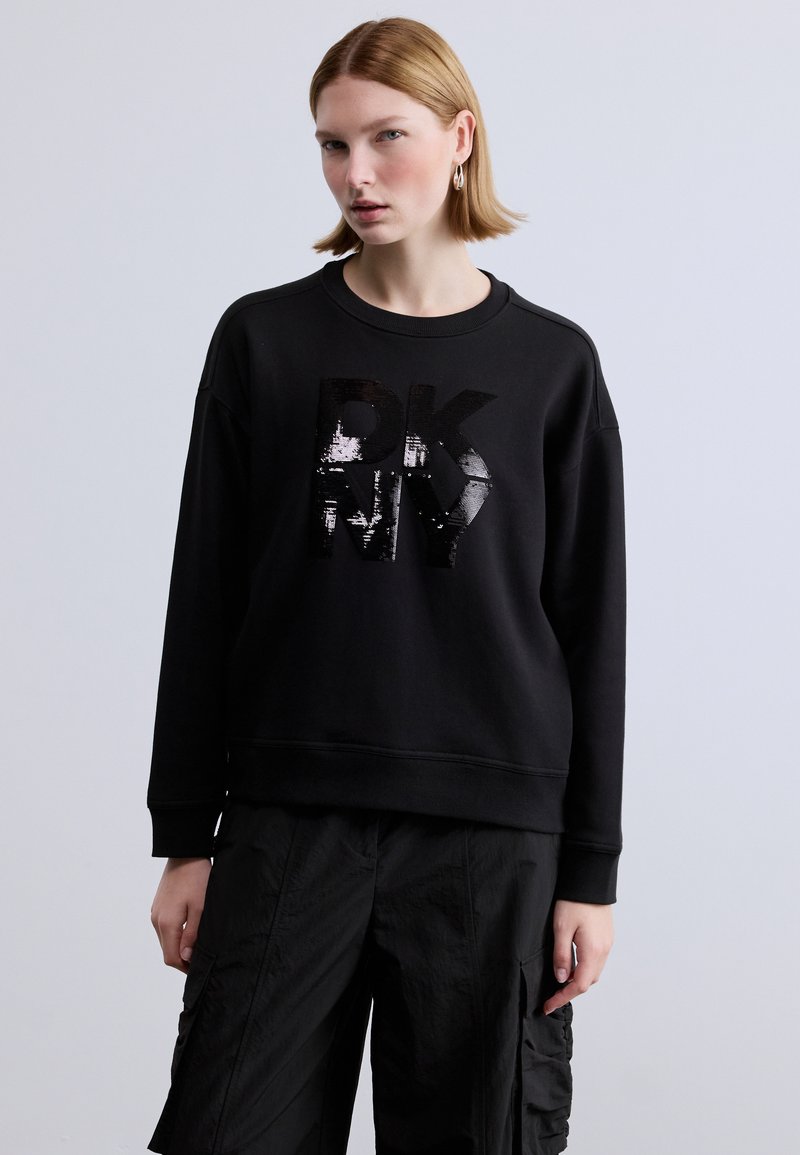 Μαύρο φούτερ με στρογγυλή λαιμόκοψη και υφή βρόχου, με γυαλιστερό μαύρο λογότυπο. Συνδυασμένο με φαρδιά μαύρη εργασιακή παντελόνα.