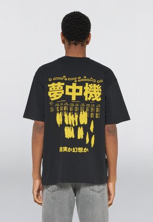 Person iført en sort t-shirt med store gule japanske tegn og abstrakte mønstre på bagsiden, kombineret med grå jeans.