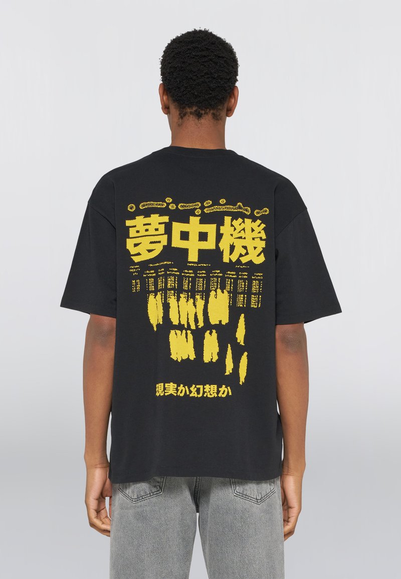 Personne portant un t-shirt noir avec de grands caractères japonais jaunes et des motifs abstraits dans le dos, associé à un jean gris.