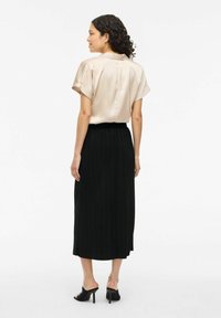 Satinblus med kort ärm i beige, parat med en svart plisserad midikjol och svarta klackmules, sedd bakifrån.