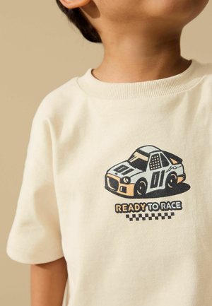 Copil purtând o cămașă bej cu o mașină de curse desenată și textul „GATA DE CURSĂ” deasupra unui design cu steag în carouri.