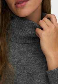 Pull en maille gris avec un col roulé haut et côtelé. Plan rapproché montrant la texture douce et les fibres naturelles. Main touchant légèrement le col.