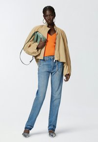 Chaqueta bomber ligera beige con mangas fruncidas, top naranja de cuello en V, vaqueros rectos azules y zapatos con estampado. Bolso verde.