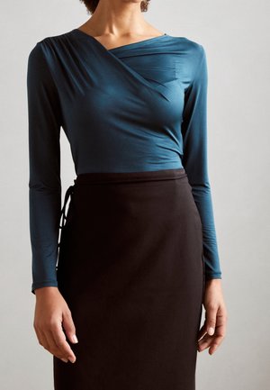 Femme portant un haut cache-cœur à manches longues de couleur sarcelle et une jupe noire taille haute avec un détail de nouage sur le côté, sur un fond uni.
