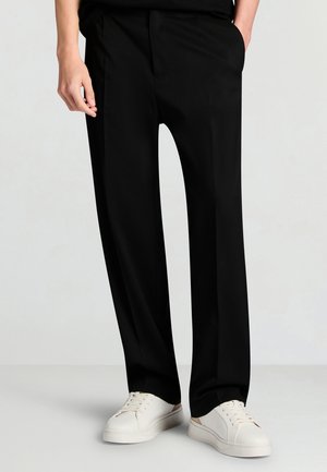 Persona che indossa pantaloni neri a gamba dritta e sneakers bianche, in piedi con una mano in tasca davanti a uno sfondo semplice.