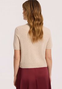 Kobieta z falującymi jasnobrązowymi włosami, ubrana w beżowy sweter z krótkim rękawem i bordową spódnicę, ukazana z tyłu na jednolitym tle.