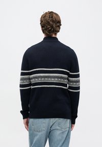 Mężczyzna z falującymi brązowymi włosami, ubrany w granatowy sweter z białymi poziomymi paskami, w połączeniu z jasnoniebieskimi dżinsami, stojący tyłem.