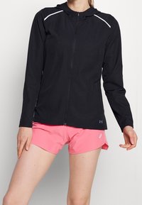 Schwarze Zip-Jacke mit Kapuze, weißen Akzentlinien auf den Schultern und leichtem Stoff. Kombiniert mit pinken Shorts, die ein sportliches Design zeigen.