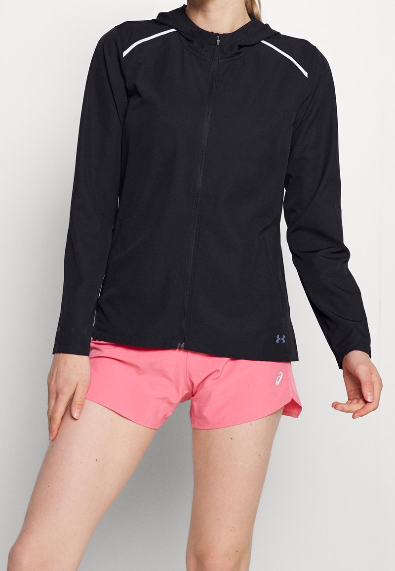 Schwarze Zip-Jacke mit Kapuze, weißen Akzentlinien auf den Schultern und leichtem Stoff. Kombiniert mit pinken Shorts, die ein sportliches Design zeigen.