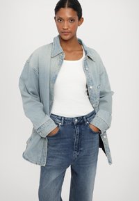 Lichtblauwe denimjack met grote zakken, knoopsluiting en laatvallende schouders, gedragen over een witte tanktop en donkeren hoge taille jeans.