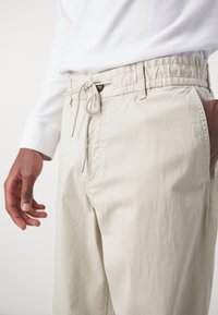 Pantalones beige de cintura elástica con cordón, bolsillos laterales y tejido texturizado. Costuras visibles y acentos metálicos en el cordón.