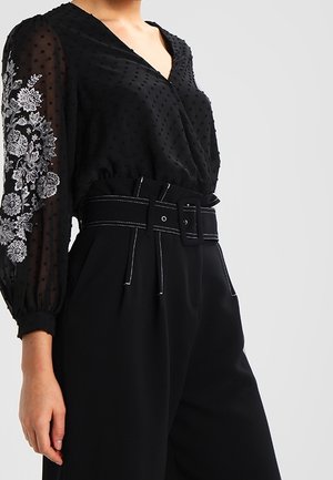 Blusa nera a pois con maniche trasparenti e ricami floreali bianchi. Abbinata a pantaloni neri a vita alta e una cintura larga.