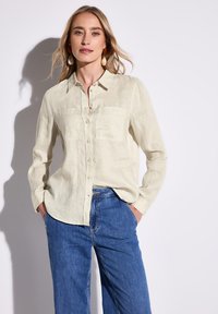 Camicia di lino beige chiaro con chiusura a bottoni, due tasche sul petto e maniche lunghe, abbinata a pantaloni in denim blu.