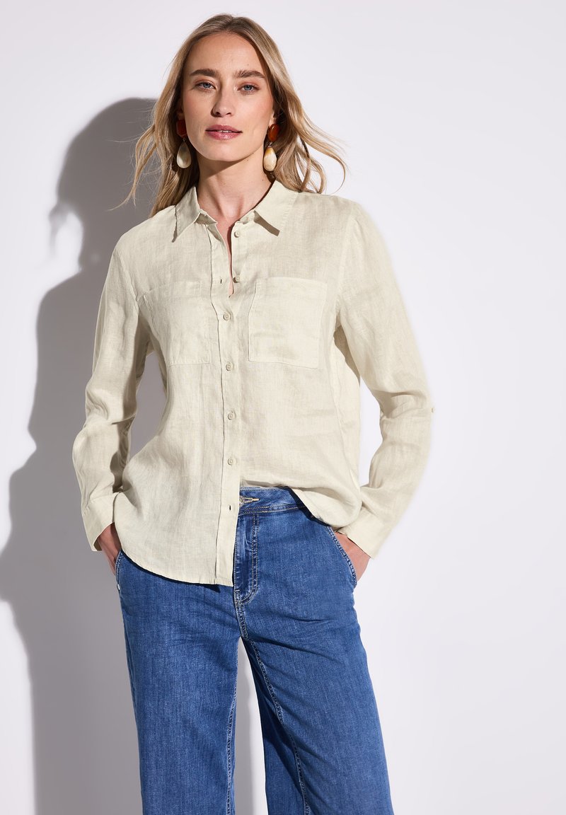 Camicia di lino beige chiaro con chiusura a bottoni, due tasche sul petto e maniche lunghe, abbinata a pantaloni in denim blu.
