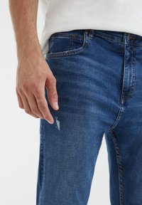 Jeans de mezclilla azul oscuro con un ligero detalle desgastado en el muslo, con un diseño clásico de cinco bolsillos y acentos de costura visibles.