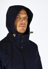 Marineblå vandtæt jakke med en justerbar hætte, fremtrædende knapper og kontrasterende brune hardwaredetaljer. Glat tekstur med minimalistisk design.
