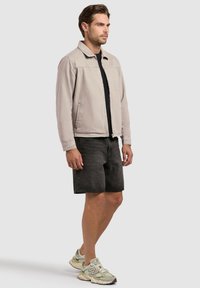 Homme portant une veste zippée beige clair, un t-shirt noir, un short en jean foncé et des baskets vert clair et beige, debout de profil.