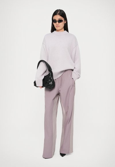 Suéter oversized roxo claro, calças altas de lavanda clara, bolsa preta e óculos de sol pretos estreitos, sobre um fundo branco.