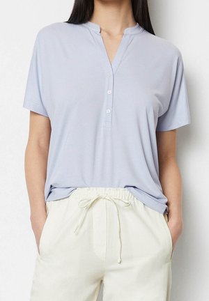 Mujer con camiseta henley de manga corta azul claro metida dentro de pantalones blancos de cintura alta con cordón, con las manos en los bolsillos.