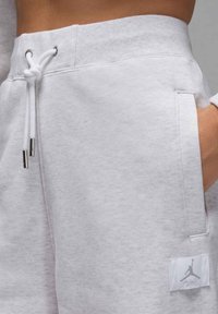 Pantalones de chándal gris claros con cintura ajustable, bolsillos laterales y un pequeño parche blanco con el logo que presenta la silueta de una figura saltando.
