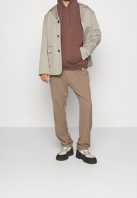 Beiger, maßgeschneiderter Blazer über einem braunen Hoodie, kombiniert mit braunen Hosen und zweifarbigen Sneakern mit schwarzen Sohlen und silbernen Hardware-Akzenten.