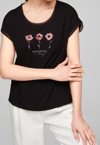 Camiseta negra de manga corta con cuello redondo. Presenta un estampado floral y el texto "HERMOSA como siempre" en colores contrastantes. Tejido suave.