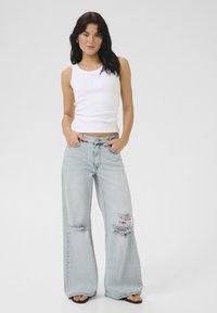 Top sin mangas blanco acanalado con un detalle bordado, combinado con jeans claros de pierna ancha y desgastados, con bordes deshilachados.