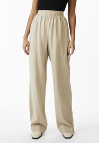 Woman wearing beige high-waisted wide-leg pants with elastic waistband and cream square-toe shoes, standing against a white background.