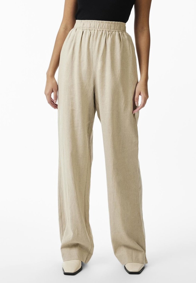 Woman wearing beige high-waisted wide-leg pants with elastic waistband and cream square-toe shoes, standing against a white background.