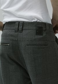 Gray patterned trousers with a textured fabric. Features include a black logo patch on the waistband and zippered back pockets.