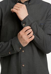 Donkergrijze visgraatblouse met knoop-omhangende kraag, bruine knopen en een klein borstzakje. Gladde textuur met een casual pasvorm.