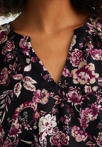 Detailní záběr na výstřih černého topu s fialovým, bílým a růžovým květinovým vzorem, který nosí osoba s středně hnědou pletí a kudrnatými vlasy.