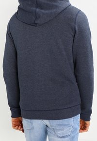 Marinblå hoodie med dragsko i huvan, ribbade muddar och nederkant, som visar ett mjukt, texturerat tyg. Enkelt mönster utan synliga logotyper.