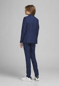 Costume marine avec un blazer ajusté et un pantalon assorti, fabriqué en tissu texturé. Associé à des baskets blanches avec des accents noirs.