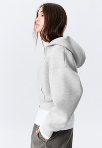 Ung kvinne med langt mørkt hår iført en lys grå glidelås-hettegenser, hvit skjorte og mørke bukser, stående i profil med hånden i lommen.