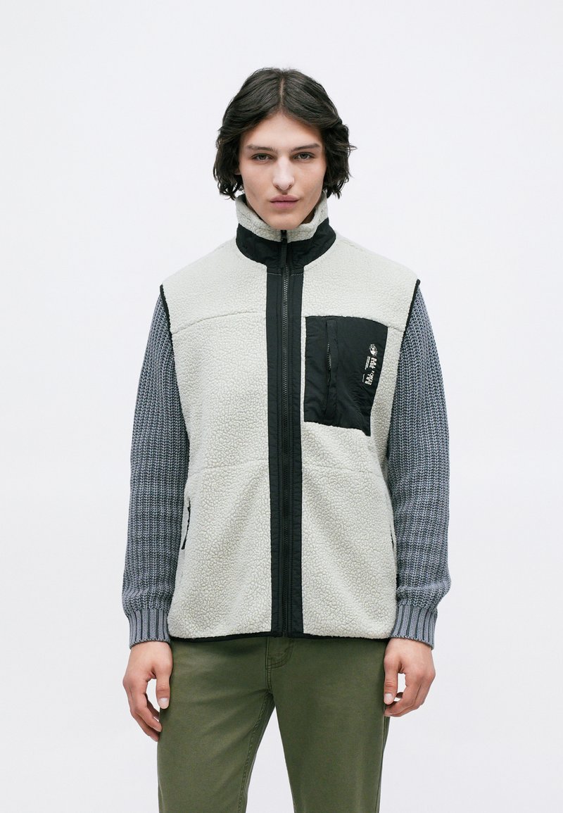 Jovem adulto vestido com um colete de fleece com fecho e bolso preto no peito, por cima de mangas de malha cinza e calças verde-oliva, em pé contra um fundo simples.