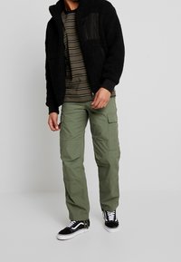 Zwarte fleece jas met een gestructureerde afwerking, olijfgroene cargobroek en zwarte sneakers met witte zolen. Het shirt heeft horizontale strepen.