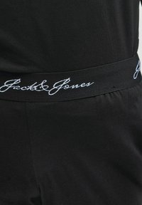 Melnā elastīgā josta ar baltu slīprakstā rakstītu "Jack & Jones" logotipu uz melnas auduma apģērba.