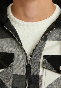 Czarna i biała kraciasta kurtka zapinana na zamek z podszewką z polaru, posiadająca wysoki kołnierz, zapięcie na zamek oraz dwie przednie kieszenie na piersi.