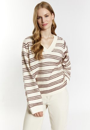 Frau mit langen, welligen Haaren, die einen cremefarbenen Pullover mit braunen horizontalen Streifen und cremefarbene Hosen trägt, steht mit einer Hand in der Tasche.