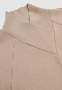 Jasno beżowa bluzka z długim rękawem z wyższym kołnierzem, miękką fakturą i subtelnym połyskiem. Szwy tworzą zarysowany wygląd.