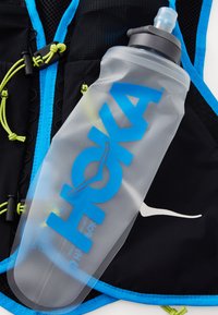 Clear plastic water bottle with a gray cap, featuring bold blue "HOKA" text. Set against a black and blue vest with neon green accents.