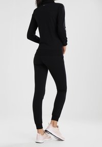 Zwart atletisch set bestaande uit een longsleeve top en nauwsluitende broek, gemaakt van gladde stof. Geaccentueerd met geribbelde boorden en gecombineerd met roze sneakers.