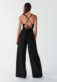 Zwarte jumpsuit met een aansluitend lijfje, gekruiste rugbandjes en een wijde pijp. Soepele textuur met een strikdetail in de taille.