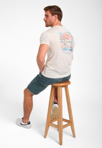 Ein weißes T-Shirt mit kurzen Ärmeln, das auf der Rückseite mit einem bunten Grafikdruck versehen ist. Dazu trägt die Person Denim-Shorts und weiße Sneakers, während sie auf einem Holzschemel sitzt.