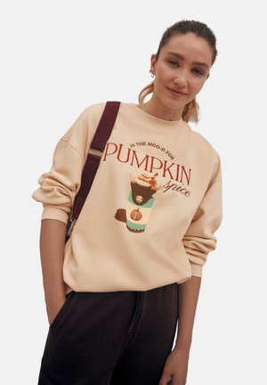 Felpa beige con la scritta "PUMPKIN spice" in rosso, accompagnata da un drink illustrato colorato guarnito con panna e accenti di arancione e marrone.