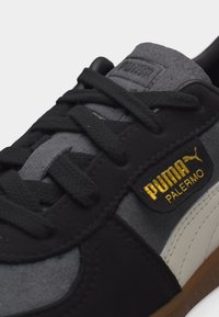 Κοντινό πλάνο μαύρου και γκρι αθλητικού παπουτσιού Puma Palermo με μαύρα κορδόνια, χρυσό λογότυπο Puma και λευκή ρίγα στο πλάι πάνω σε καφέ σόλα.